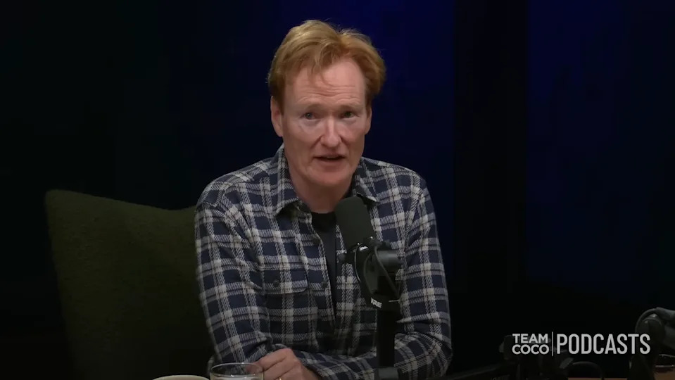 Man wearing a plaid shirt speaking into a microphone on a podcast. "Team Coco Podcasts" logo visible