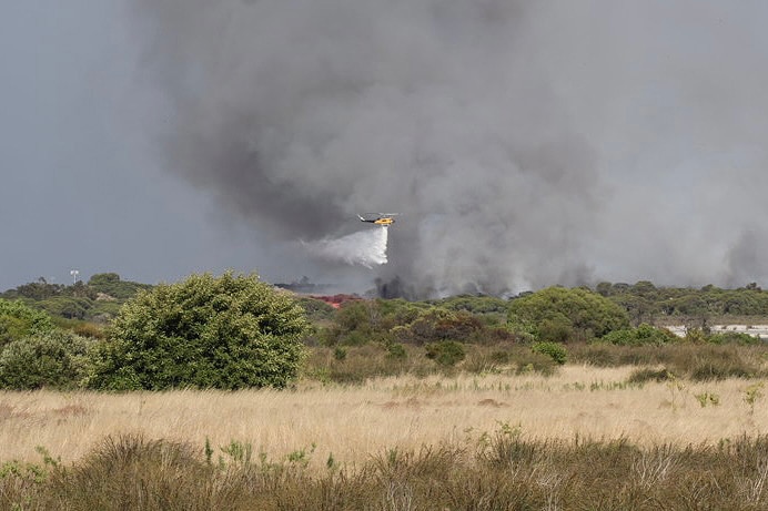 A helicopter water bomber flies over a fire in bushland, dropping a load of water on the flames.