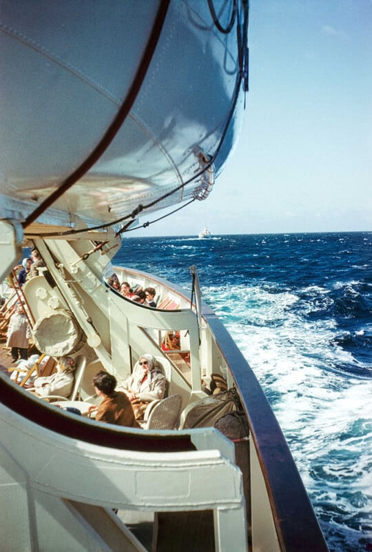 Passengers sit on deck chairs under a large lifeboat on the side of a ship, looking out at the blue ocean as the vessel moves forward, leaving a foamy wake behind. The sky is clear and bright.