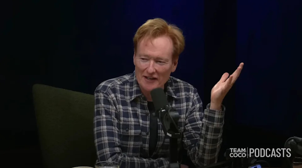 A person sits at a podcast mic, gesturing with one hand, wearing a plaid shirt. Team Coco Podcasts logo in the corner