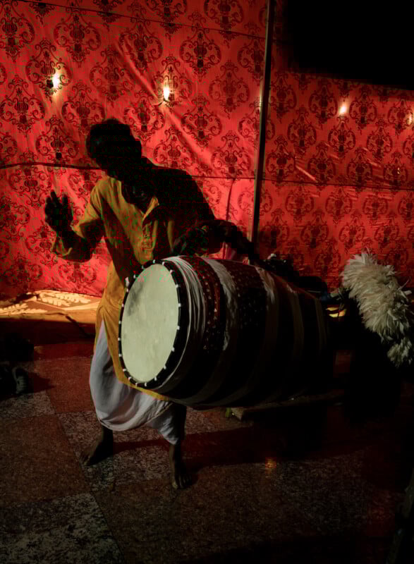 A person in traditional attire plays a large drum in front of a red, patterned backdrop, with warm lights casting dramatic shadows and highlighting the festive atmosphere.
