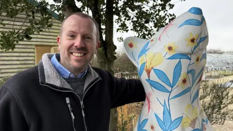 Amber Macey/BBC Ian Ridpath - a smiling man wearing a dark-coloured jacket standing next to a colourful rat sculpture. The artwork features blue and yellow flowers and is looking upwards.