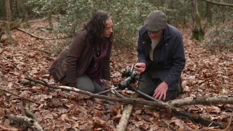 Barry Webb kneels on the forest floor amongst the leaves and branches looking into his camera, and explaining the process to reporter Victoria Cook who is kneeling next to him