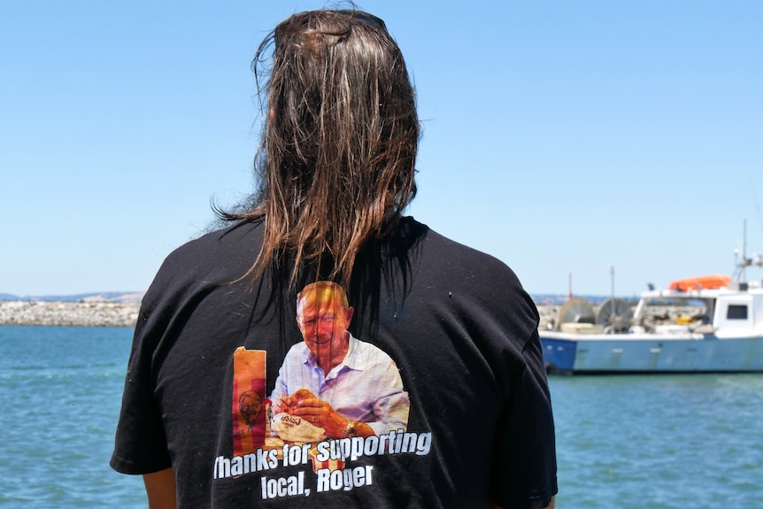 A man wearing a t-shirt saying 'Thanks for supporting local, Roger' looks at the ocean