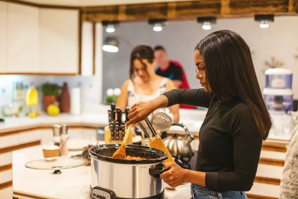 A woman stirs food in a slow cooker in a modern kitchen, with others in the background, suggesting a casual home cooking scene