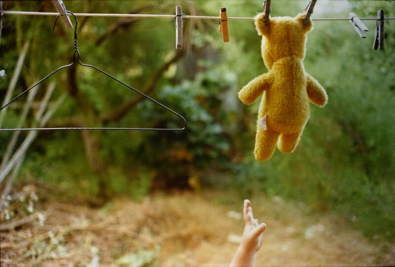 A yellow teddy bear hangs from a clothesline outdoors next to a metal hanger, with a child's hand reaching up toward it. The background is blurred greenery.
