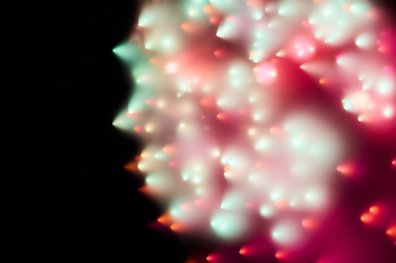 Abstract image with soft, colorful lights in shades of pink, red, white, and green, creating a blurred, glowing effect against a dark black background. The lights form a profile-like shape on the left side.