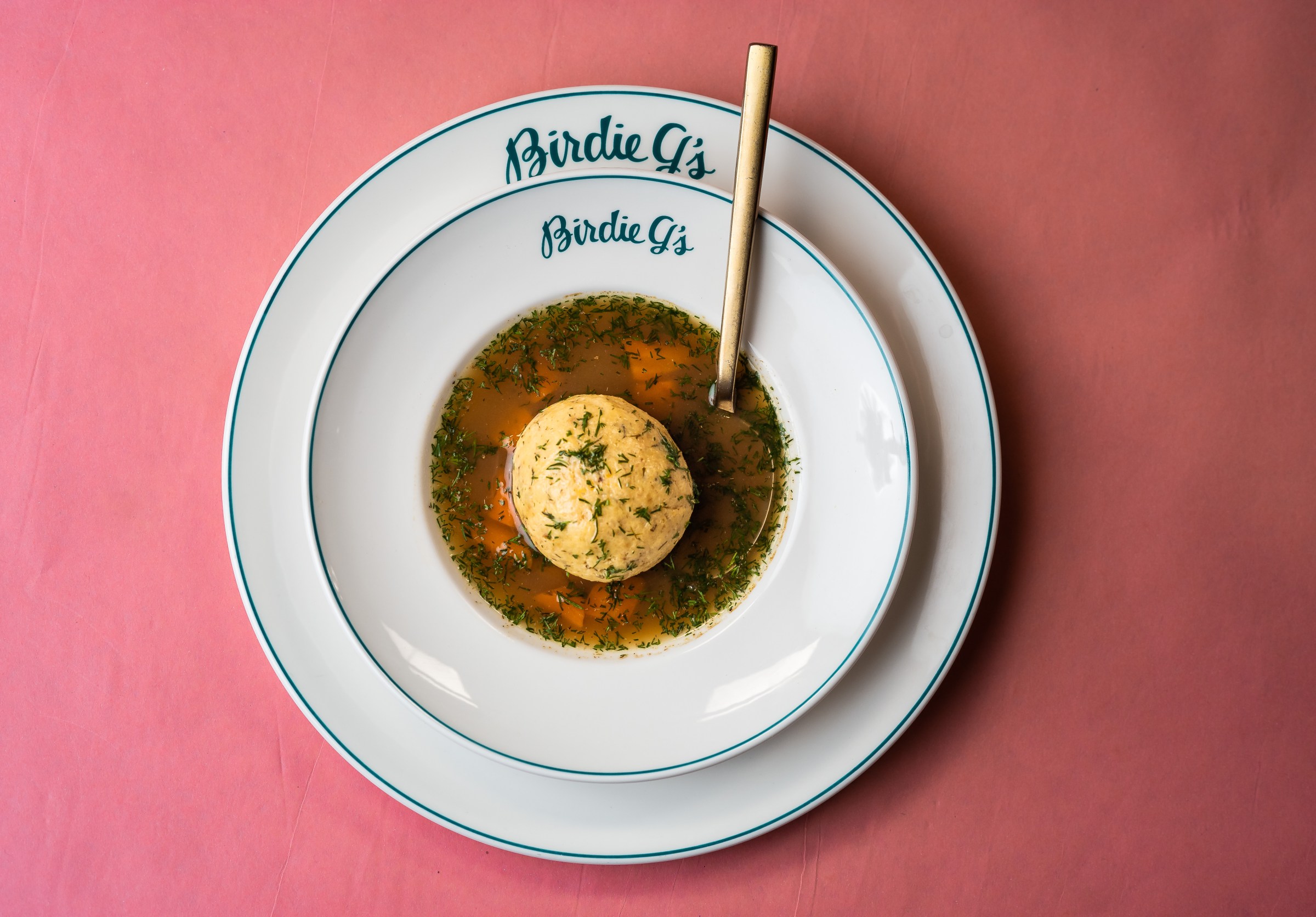 A white bowl filled with matzo ball soup atop a white plate inscribed with Birdie G’s.