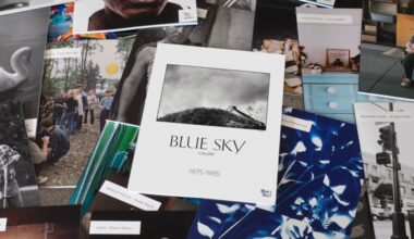 A variety of photography exhibition flyers and postcards are spread out, with a central catalog titled "Blue Sky Gallery 1975-1985" featuring a black-and-white photo of a hill with a house at the top.