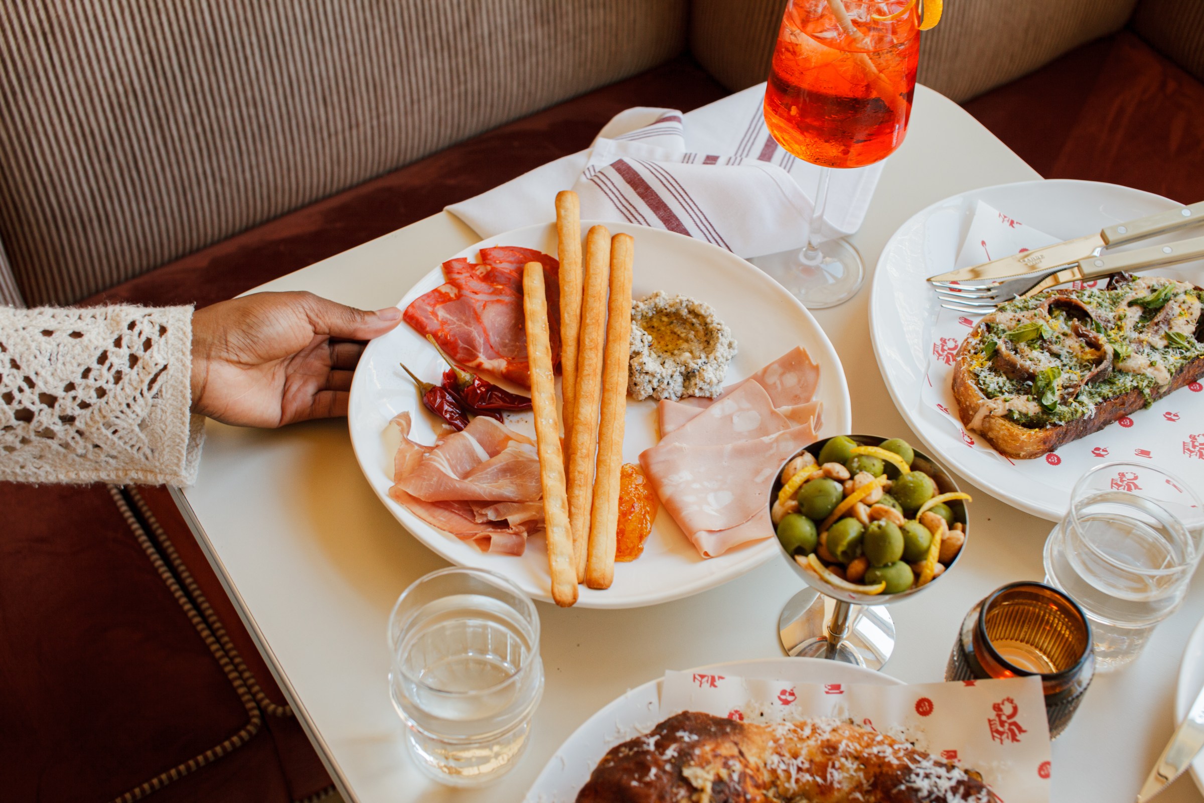 A hand holding a plate of meats, spreads, and breadsticks on a table filled with dishes and drinks.
