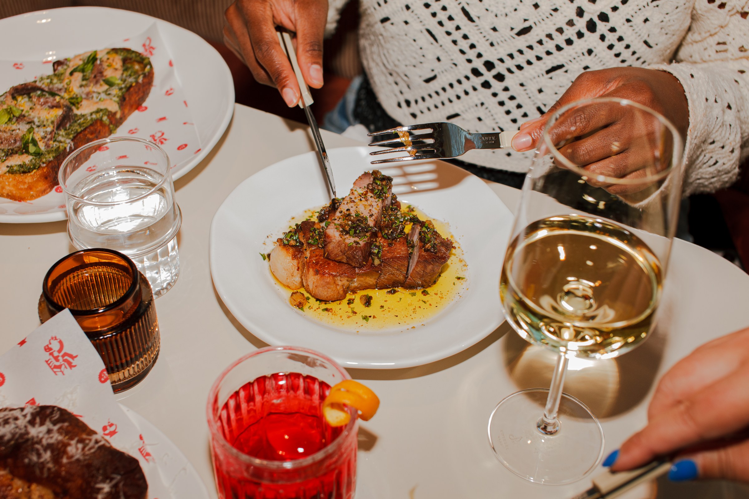 A person slicing into a piece of pork collar on a table filled with dishes and drinks.