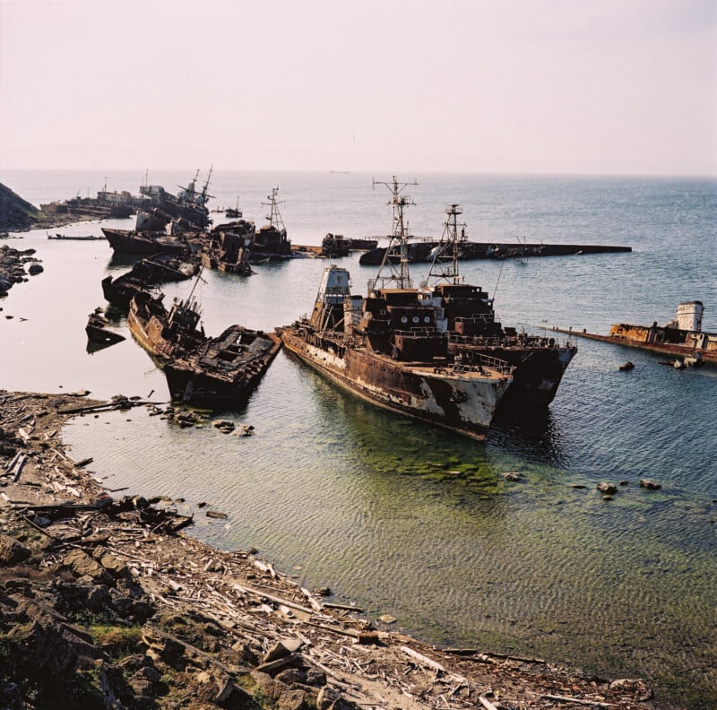 Abandoned and rusting ships lie partially submerged in shallow coastal waters near a rocky shoreline, with more shipwrecks visible further in the sea under a hazy sky.
