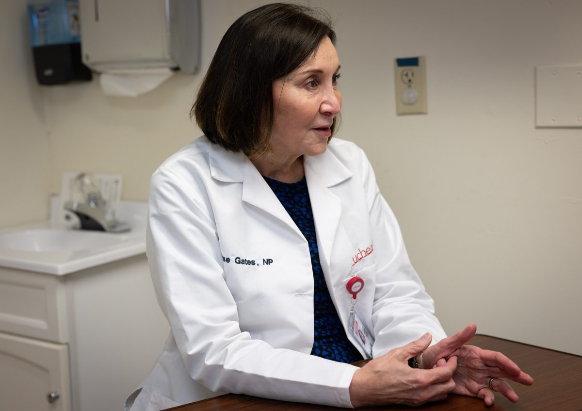 A woman in a white medical coat sits at a desk and speaks, gesturing with her hands in a clinical office setting.