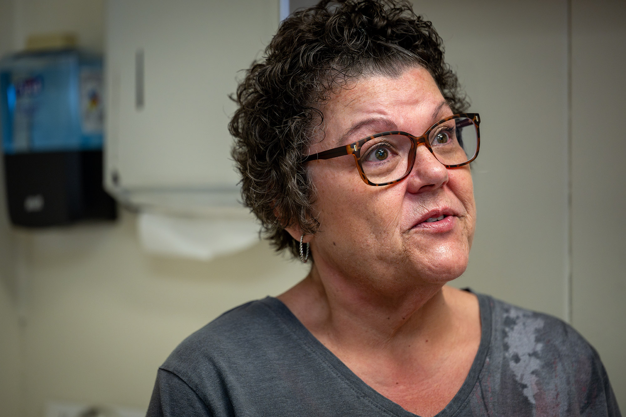 A woman with short curly hair and glasses, wearing a gray shirt, stands indoors in a room with medical supplies visible in the background.