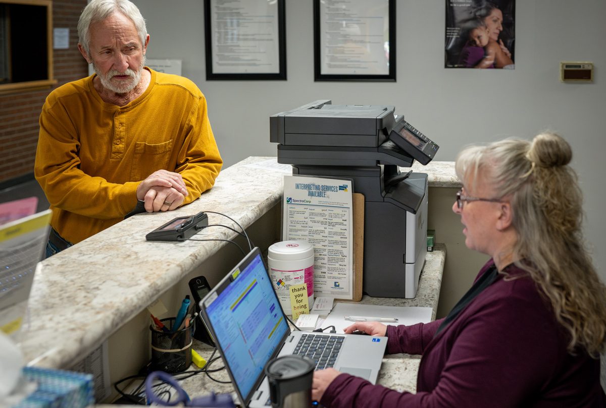 An older man stands at a reception counter while a woman types on a laptop behind the desk in an office setting.