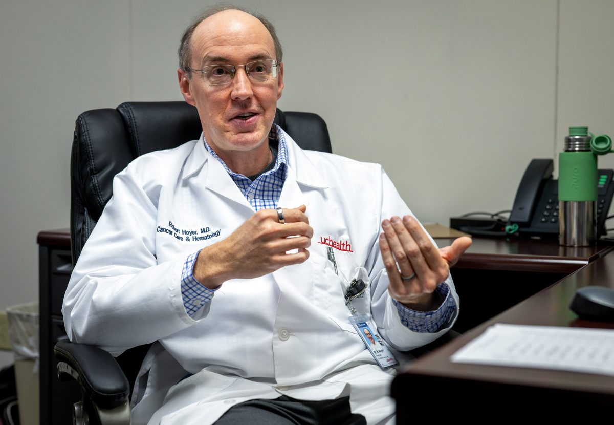 A doctor in a white lab coat sits at a desk, gesturing with his hands while speaking, with office supplies and a phone visible in the background.