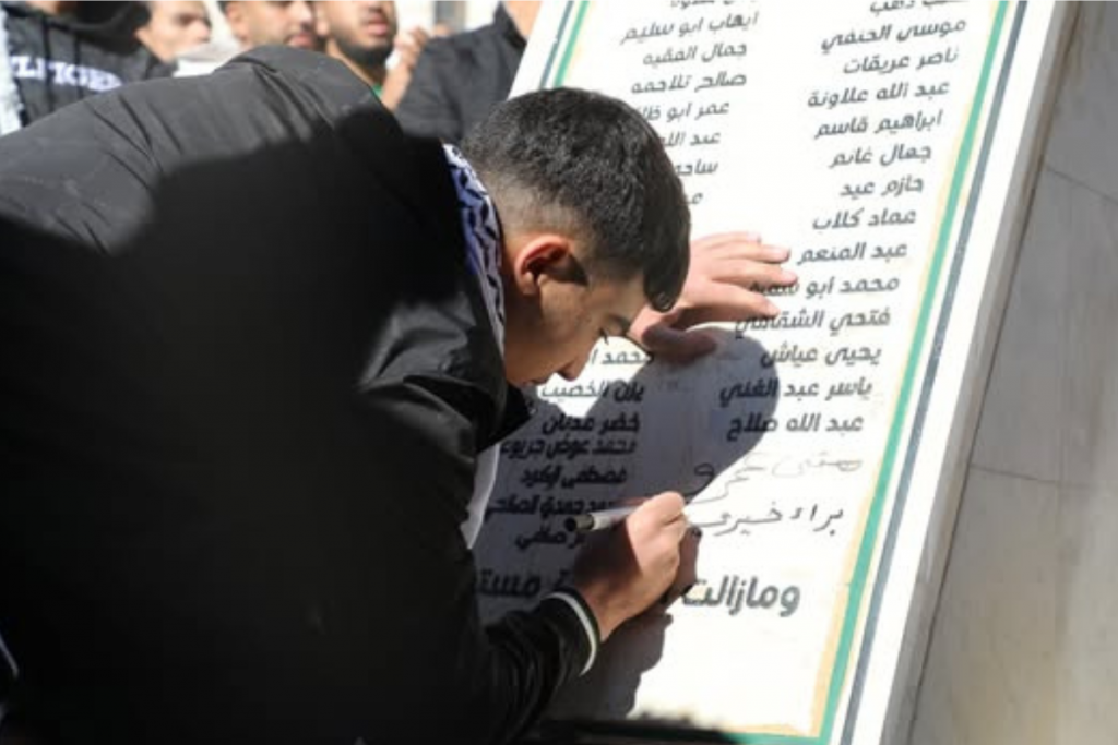 A young man writes on a memorial stone inscribed with names. People gather around him during a solemn commemoration.