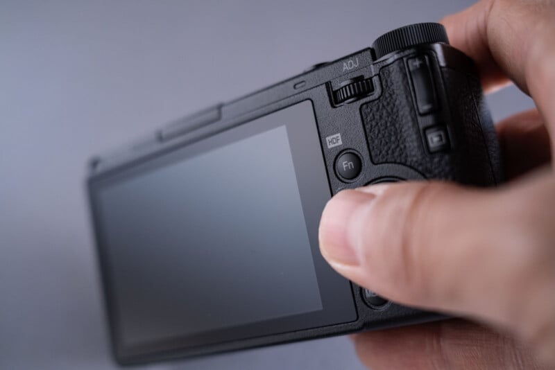 A close-up of a person's hand holding a digital camera, focusing on the camera's back controls and LCD screen with a neutral gray background.