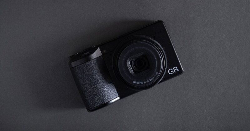 A black compact digital camera with a large lens, labeled "GR," placed on a dark gray surface at a slight angle.