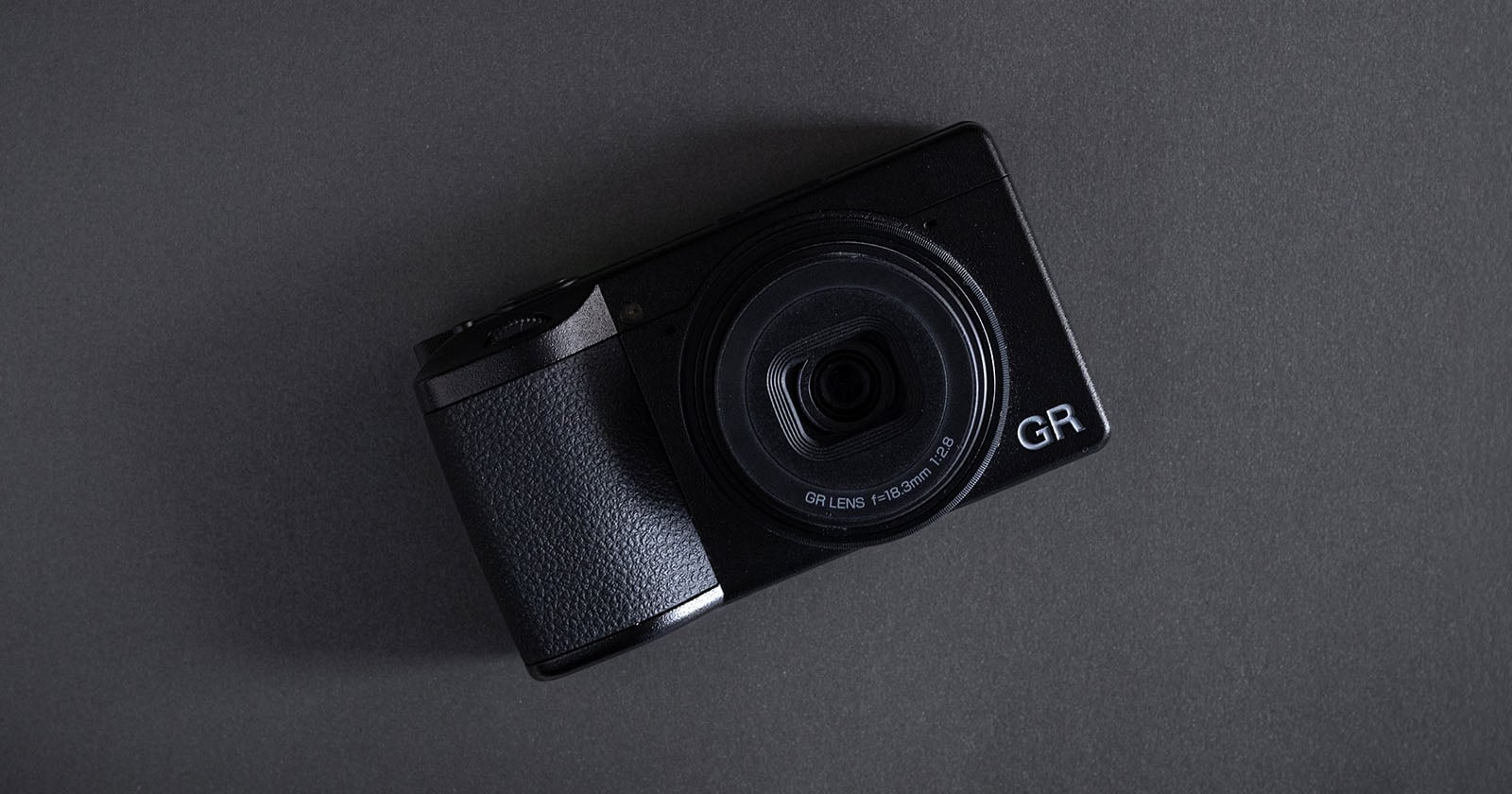 A black compact digital camera with a large lens, labeled "GR," placed on a dark gray surface at a slight angle.