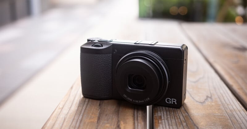 A compact black digital camera with a protruding lens sits on a wooden table outdoors, with a blurred background of greenery and pavement.