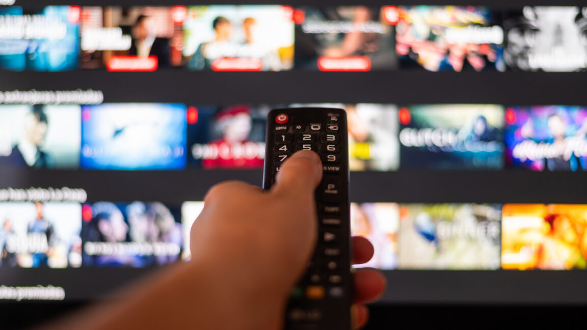 Hand Using A Television Remote Control in front of a TV with a streaming app on display and blurred in the background.