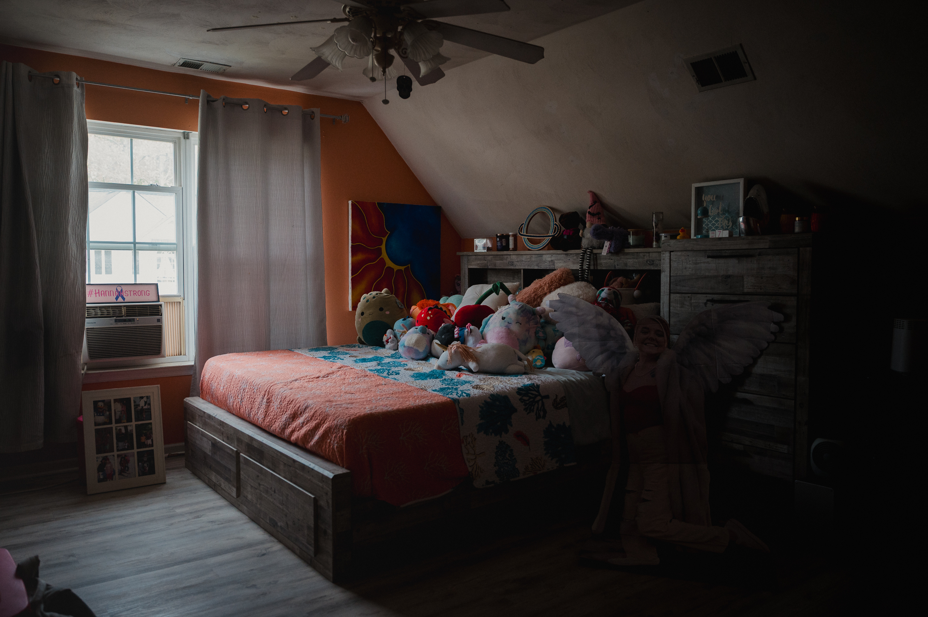 A bed sits at the center of a small bedroom with an orange wall. The bed is covered in stuffed animals.