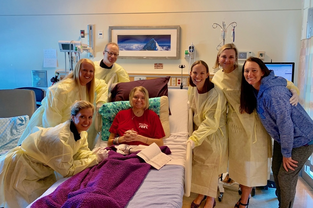 A young blond girl sits in a hospital bed propped up with pillows. She is surrounded by people wearing scrubs.
