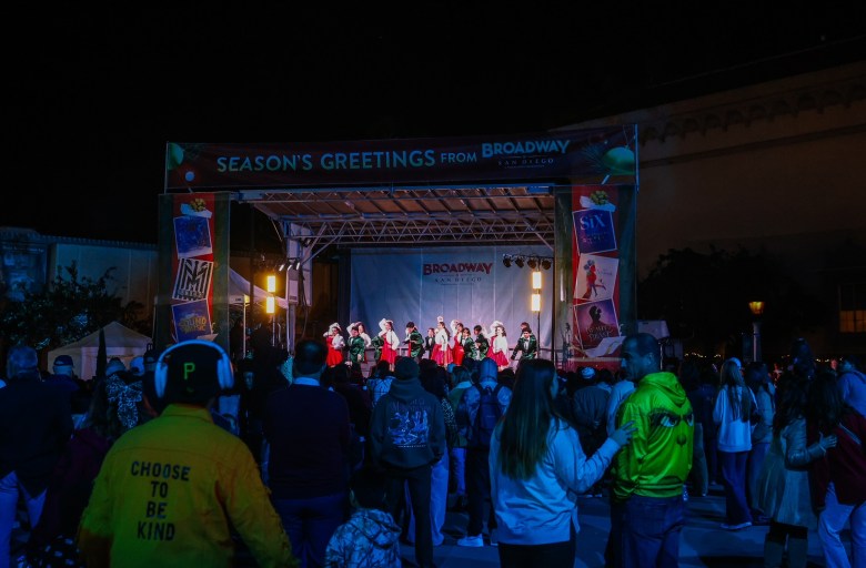 A crowd is assembled at night in front of a stage with the sign "Season's Greetings from Broadway San Diego."