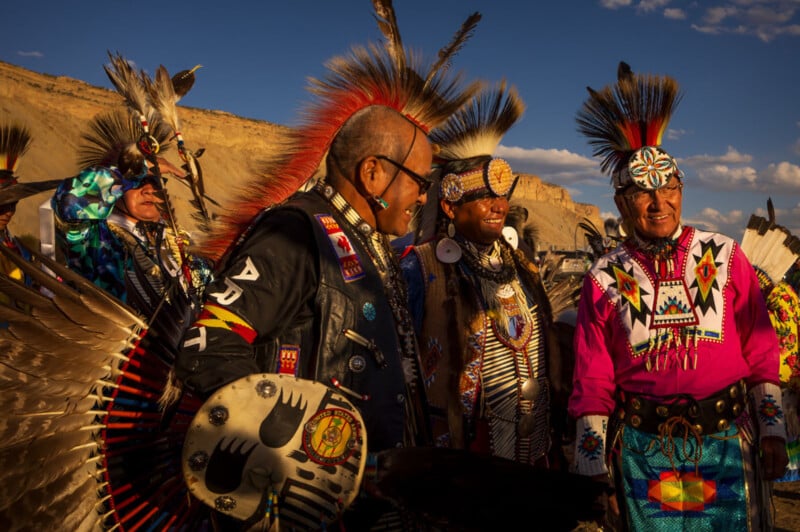 Four Indigenous men wearing colorful, traditional regalia and feathered headdresses stand together outdoors, smiling and talking, with a natural, sunlit landscape and blue sky in the background.
