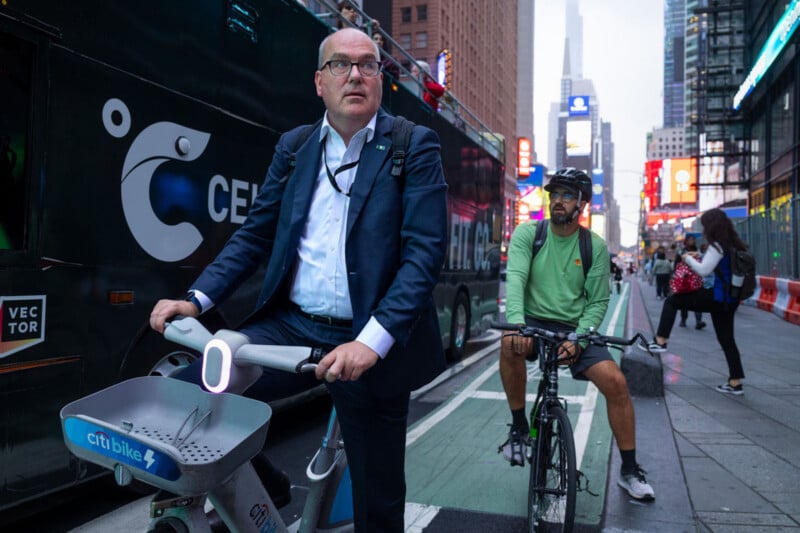 A man in a suit rides a Citi Bike next to a man in casual clothes on a bicycle in a city bike lane surrounded by tall buildings and bright lights in the background.