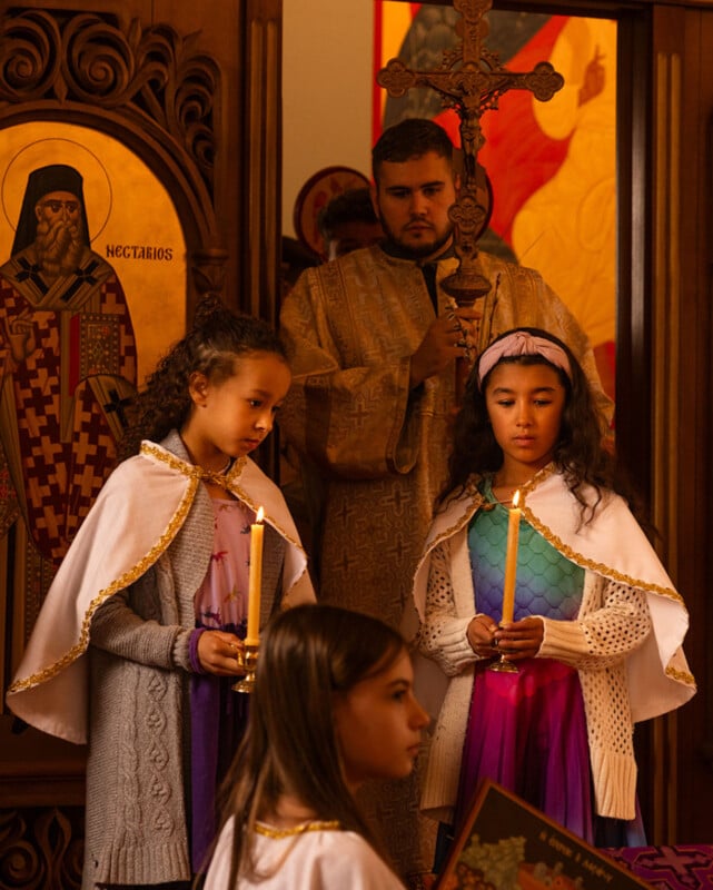 Two young girls in costumes and white capes hold candles in a church, accompanied by a man in religious attire holding a cross. Religious icons and warm lighting appear in the background.