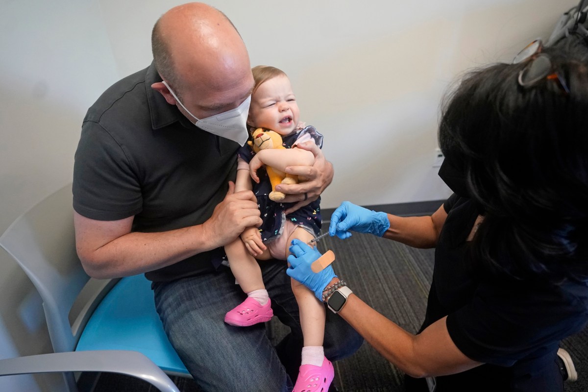 A man holds a crying toddler while a healthcare worker applies a bandage to the child's leg after a vaccination.