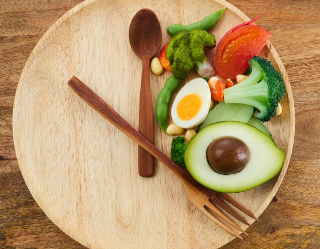 Food on a wooden plate in one section with cutlery placed to like hands on a clock