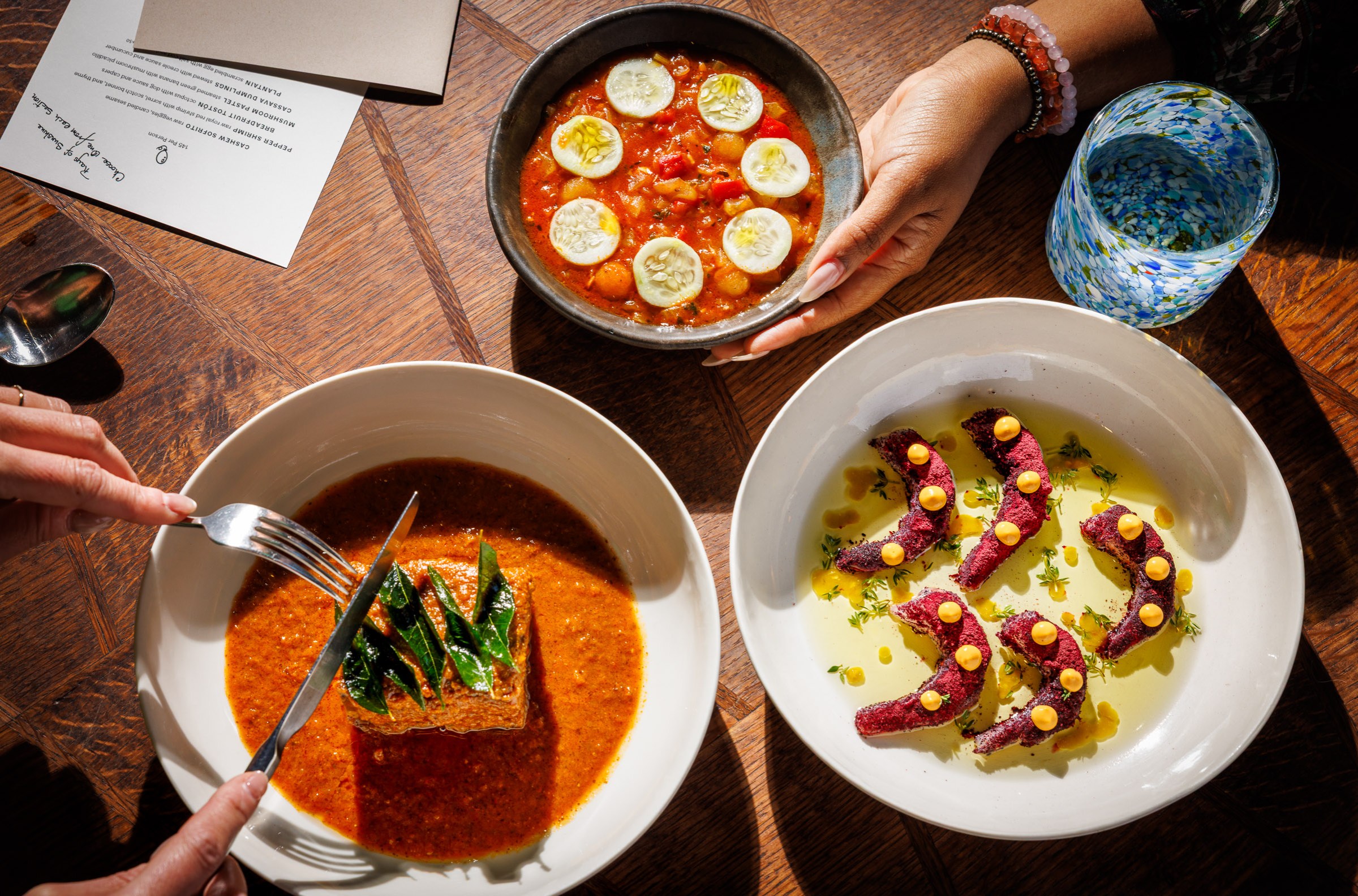 Cassava dumplings, raw shrimp dusted with sorrel, and a hunk of goat shoulder in three white bowls on a wooden table.