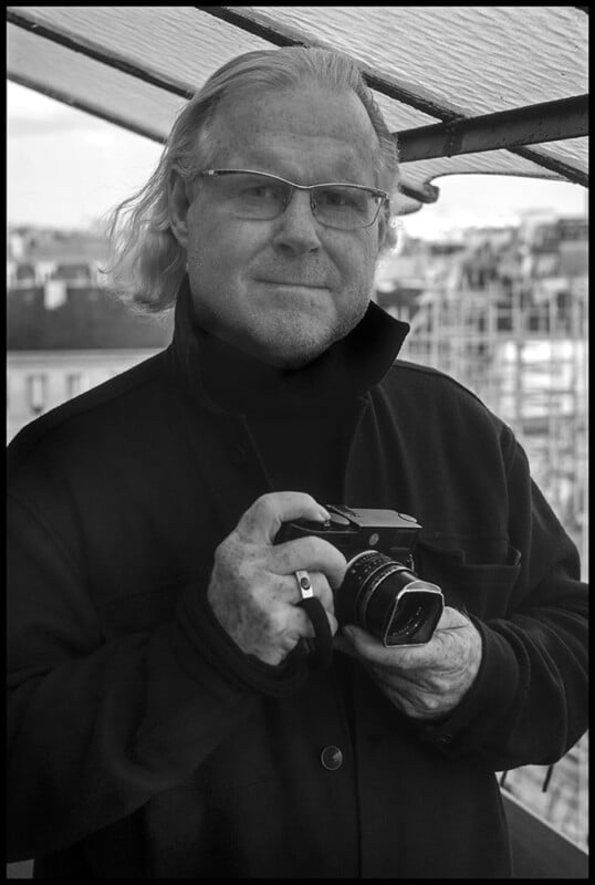 A man with light hair and glasses, wearing a dark coat, stands outdoors under a canopy and holds a camera in his hands. The background shows buildings and scaffolding. The image is in black and white.