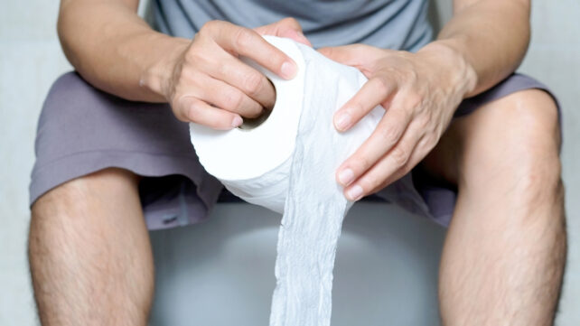 A man sitting on a toilet, fully clothed, holding a roll of toilet paper. To illustrate a connection between bowel movement frequency and overall health