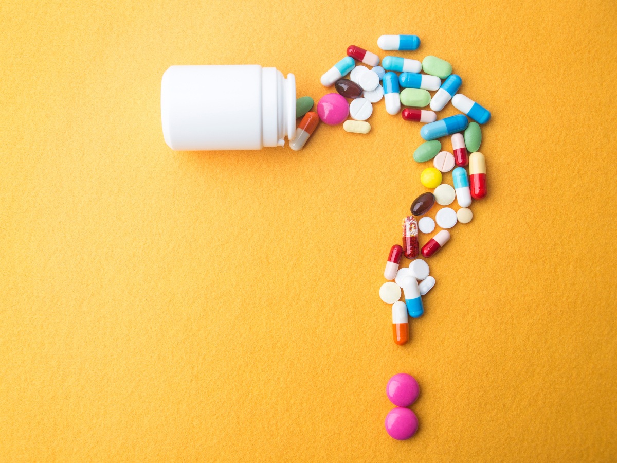 Various colourful medication tablets arranged in the shape of a question mark with a small plastic bottle at the top, against a yellow background.