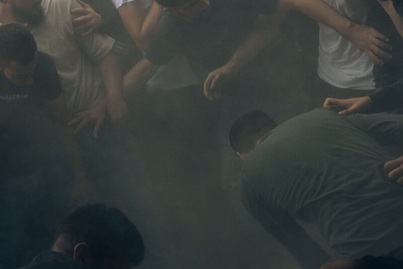 A group of people stand closely together in a smoky or dusty environment, some reaching out with their hands, their faces partly obscured by the haze.