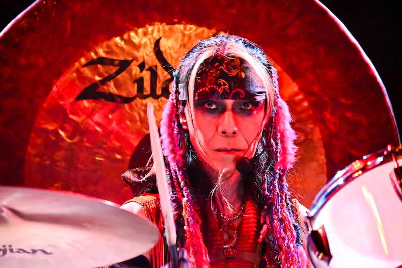 Atsuo, wearing a bandana, colorful strad briad and smoky eye makeup, starts in front of the audience while playing drums.