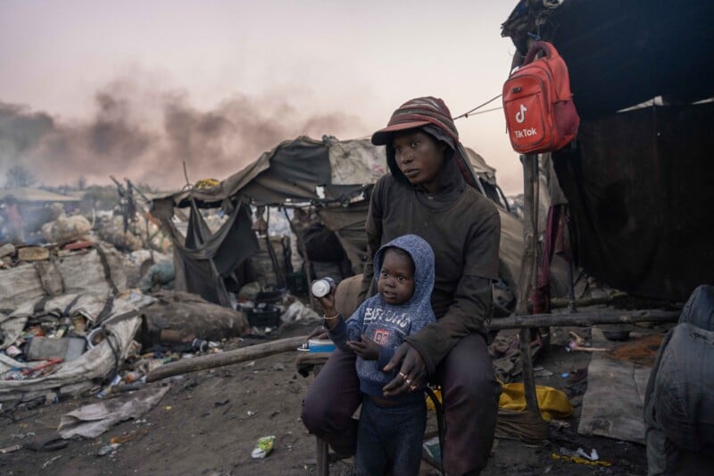 A person sits with a small child on their lap in a makeshift camp surrounded by debris and smoke. Tents and shelters made from tarps and fabric are visible, with a red backpack hanging nearby. The scene appears tense and somber.
