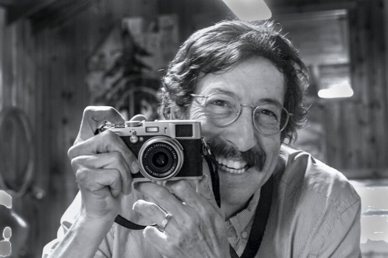 A smiling man with glasses and a mustache holds a vintage camera up to his face indoors. The black-and-white photo captures a joyful, candid moment.