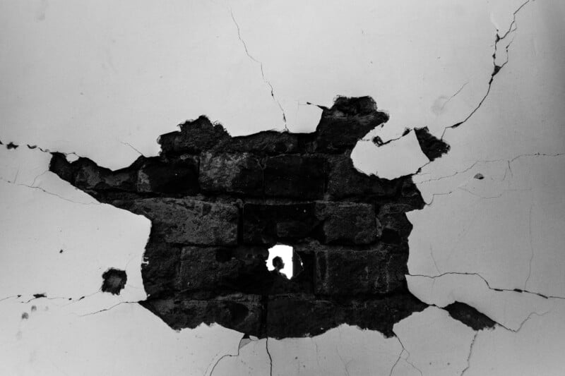 A large hole with cracks surrounds exposed bricks in a wall; through a small opening in the center, a silhouette of a person is visible against bright light in the background.