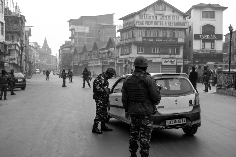 Armed soldiers in camouflage uniforms stop and inspect a car on an empty city street lined with commercial buildings and shops in the background; the scene appears tense and foggy.