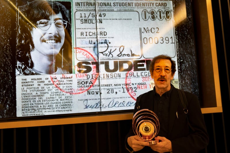 A man stands holding an award in front of a large projection of an old International Student Identity Card featuring his younger self, labeled “Richard” from York College, with a black-and-white portrait and official stamps.