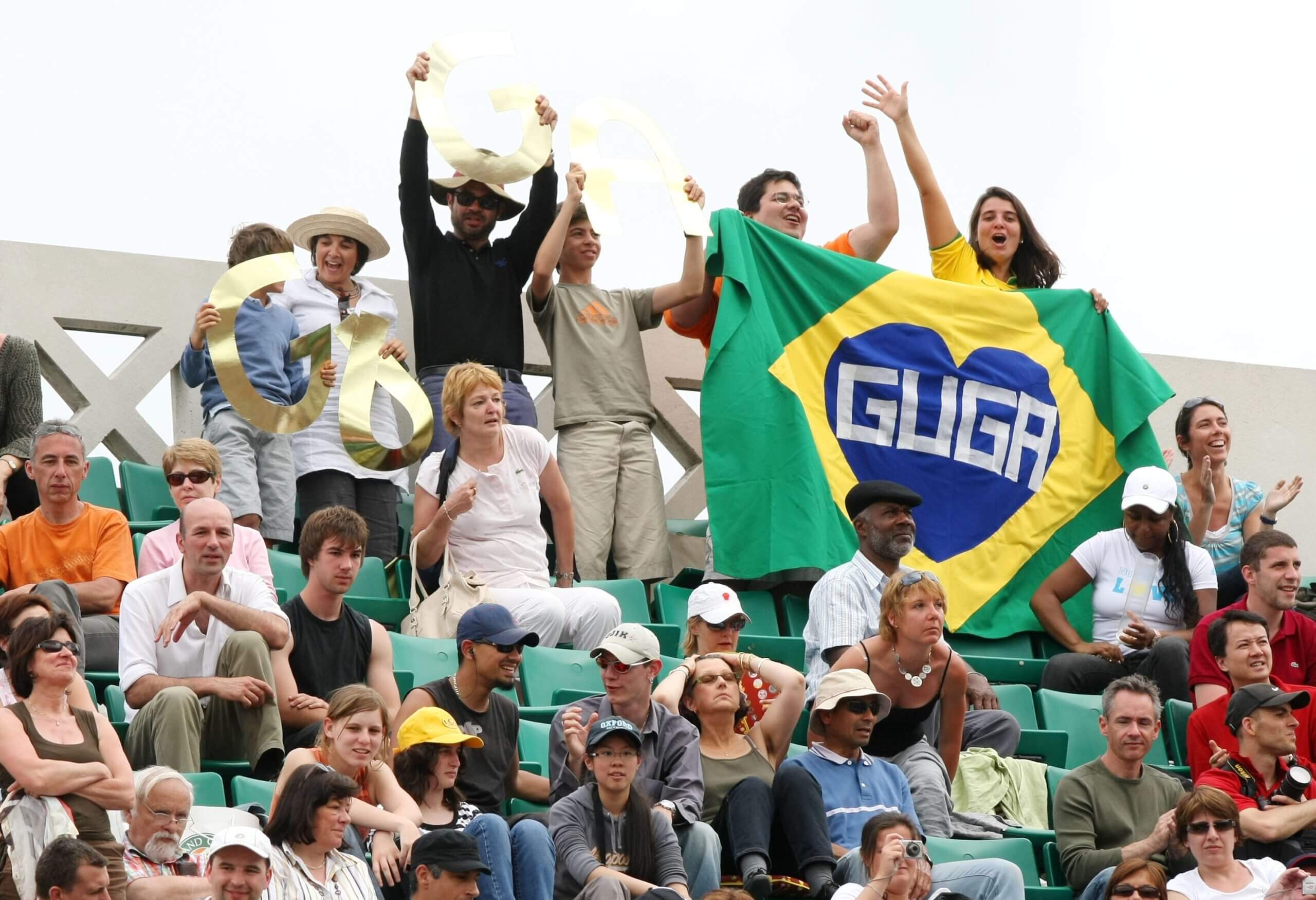 Fans at a tennis match stand holding a Brazilian flag with the word “Guga” in the center in white capital letters.