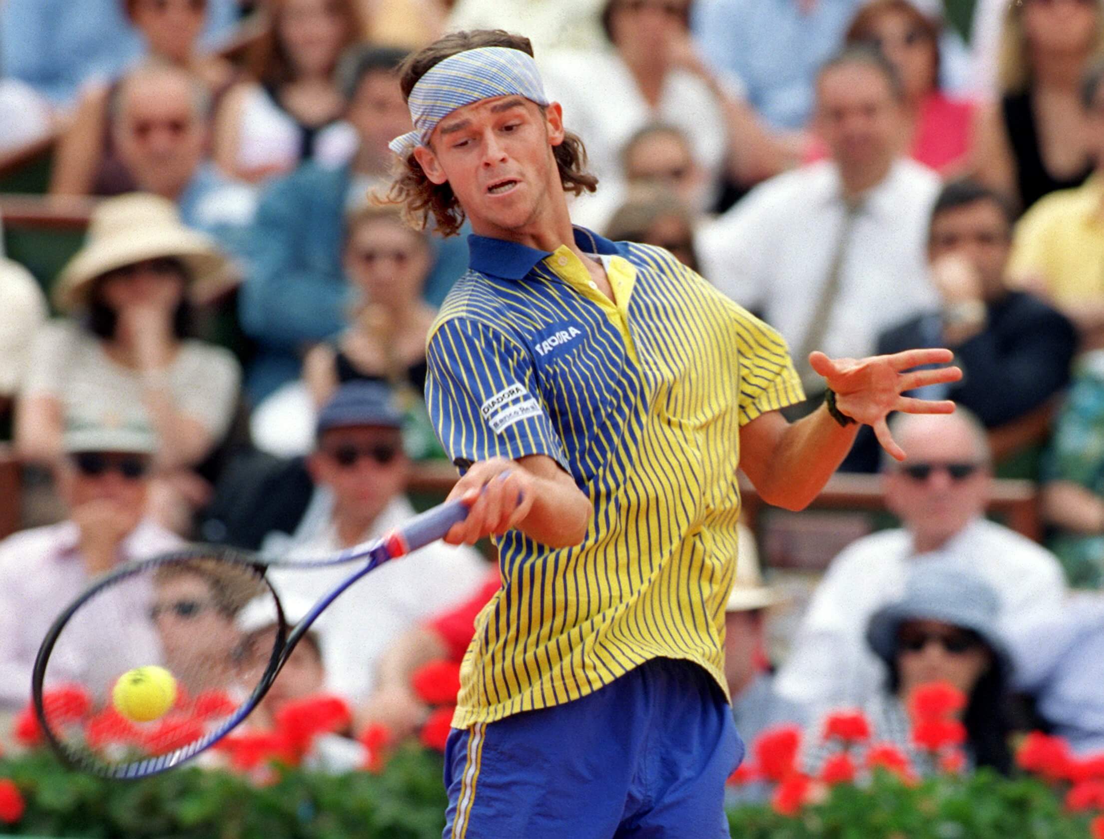 Gustavo Kuerten wears a blue-and-yellow-striped shirt and a headband as he whips a topspin forehand with his eye on the ball on the strings of his racket.