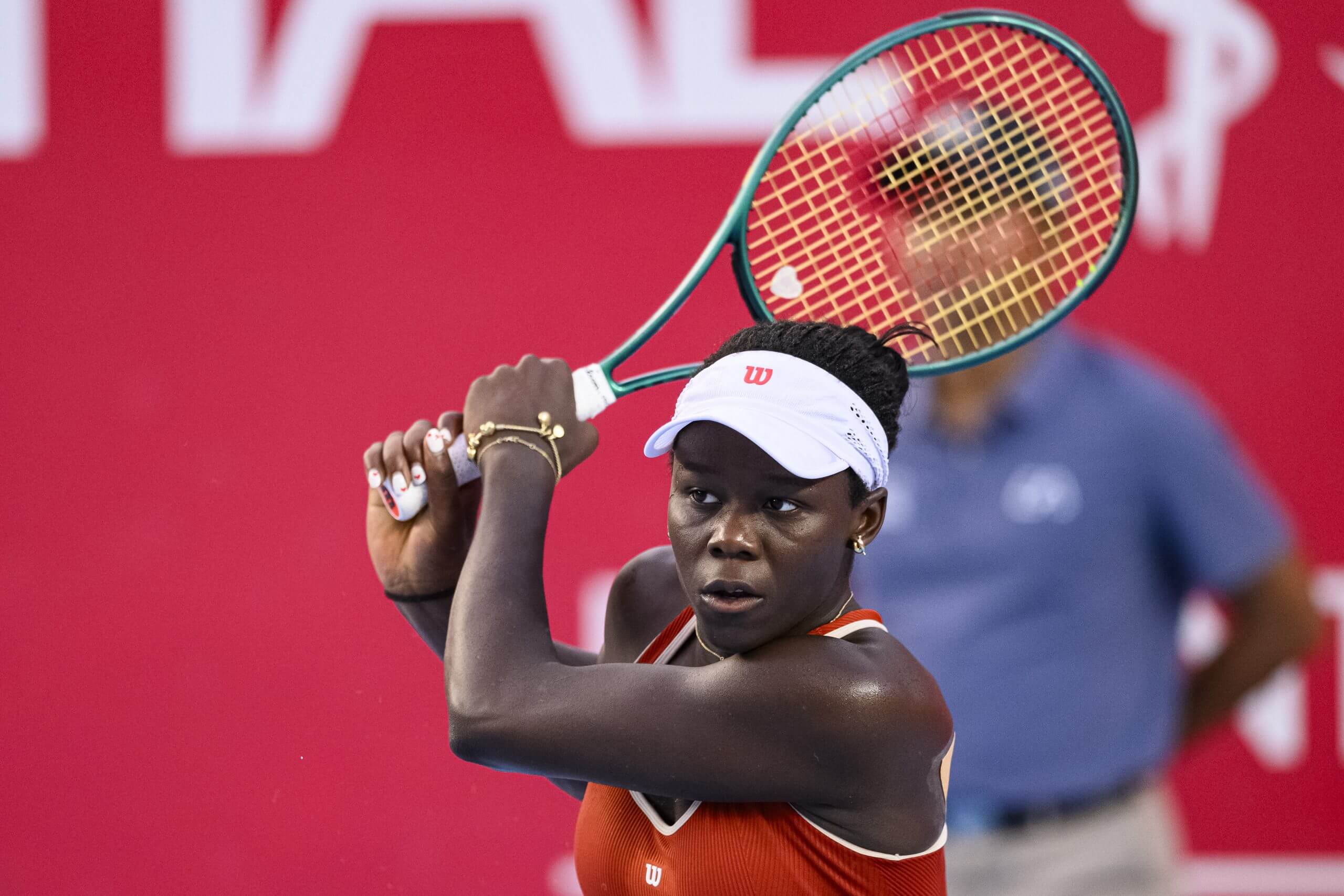 Victoria Mboko follows through on a two-handed backhand with her racket above her head, as she wears a white visor.