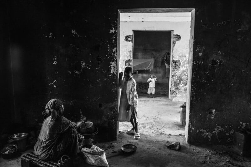 A black and white photo shows a woman sitting on the floor preparing food near a wall, while a child stands in a doorway and another child stands farther back in a separate, sparsely furnished room.