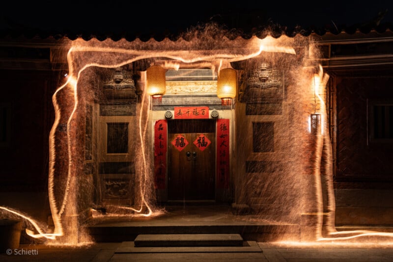 A traditional Chinese entrance with red lanterns and calligraphy banners, illuminated at night. Golden light trails swirl around, creating glowing patterns in front of the doorway.